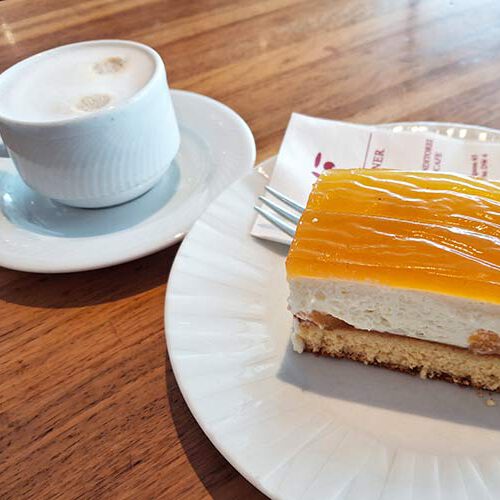 Eine Tasse Melange und daneben auf einem Teller eine Pfirsich Joghurt Schnitte.